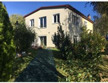 Zweistöckiges Wohnhaus mit hellgelber Fassade und roten Dachrändern, umgeben von einem Garten mit Bäumen, Sträuchern und Rasen. Ein mit grünem Material ausgelegter Weg führt durch den Garten, links steht eine schwarze Laterne.