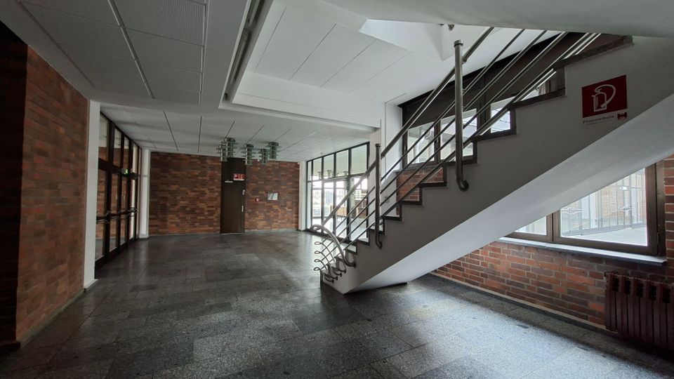 Innenbereich eines Treppenhauses in einem Gebäude mit Backsteinwänden, dunkelgrauem Steinboden und weißer Decke. Eine Treppe mit Metallgeländer führt nach oben, daneben große Fensterfronten und Glastüren entlang des Flurs.