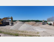 Eine Baustelle mit mehreren Schotter- und Erdhaufen, einem gelben Bagger links im Bild und Baugeräten sowie Containern im Hintergrund. Im Vordergrund sind unbefestigte Wege, und im Hintergrund ist eine Baumreihe unter einem klaren blauen Himmel sichtbar.