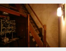 Treppenhaus mit einer einfachen Holztreppe, die an einer grob verputzten Wand entlangführt. Links im Bild ist ein mechanischer Apparat mit Zahnrädern und Riemen sichtbar, während eine Wandlampe den Raum schummrig beleuchtet.