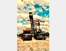 Großes Industriemotiv mit einem alten, rostigen Fahrzeug und einem grünen Lastwagen im Vordergrund, umgeben von sandigem Boden. Im Hintergrund steht ein großer industrieller Kran unter einem sonnigen Himmel mit Wolken.