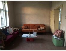 Ein spärlich eingerichteter Wohnraum mit schlichten, hellen Wänden. Zu sehen sind ein rotes und ein orangefarbenes Sofa, ein kleiner weißer Tisch mit Gegenständen darauf und ein Fenster links, das Tageslicht hereinlässt.