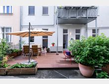 Ein moderner Innenhof mit einer Holzterrasse, ausgestattet mit einem großen Sonnenschirm, Gartenmöbeln und bepflanzten Hochbeeten. Im Hintergrund ist eine helle Hausfassade mit Fenstern, einem Balkon und Pflanzen zu sehen.