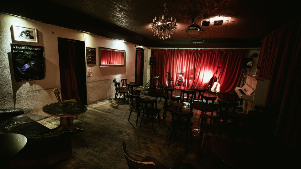 Dimly lit small performance venue with wooden floors, café-style tables and chairs, and a low stage backed by red curtains. A chandelier hangs from the ceiling, and the stage area includes microphones, an acoustic guitar, and an upright piano.