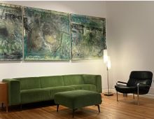 Wohnzimmer mit Holzboden, einer grünen Samtcouch mit passendem Hocker, einem schwarzen Ledersessel und einer modernen Stehlampe. An der weißen Wand hängen drei große, abstrakte Gemälde in grünen und bläulichen Farbtönen.