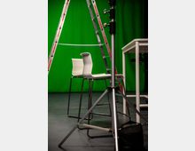 Ein Studio mit grünem Hintergrund (Greenscreen), zwei weißen Barhockern, einer offenstehenden Leiter und einem Stativ im Vordergrund. Auf der rechten Seite steht ein weißer Tisch.
