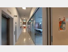 Ein moderner Innenflur mit glatten, hellen Böden, auf der rechten Seite verglaste Seminarraumtüren mit Tischen und Stühlen im Inneren. Links ist ein Fahrstuhl sichtbar, und am Ende des Flurs führt eine Öffnung in einen Bereich mit Büchergestellen. Ein Plakat mit der Aufschrift „Wir stärken Demokratie“ hängt rechts an der Wand.