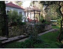 Ein idyllischer Garten mit Rasen, Bäumen und Sträuchern umgibt ein weißes Gebäude mit rotem Ziegeldach. Im Vordergrund führt ein gepflasterter Weg entlang einer kleinen Steinterrasse mit Rankgerüst und einem Sitzbereich mit Gartentisch und Stühlen.