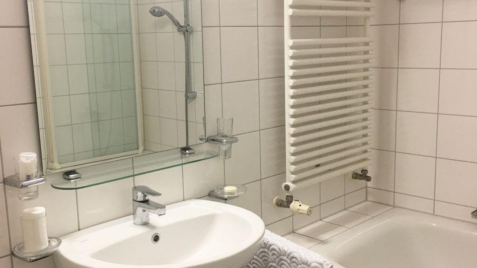 Modernes, weiß gefliestes Badezimmer mit Waschbecken und Wandspiegel samt Glasablage sowie einer Badewanne mit Duscharmatur. An der Wand neben der Wanne befindet sich ein großer, vertikaler Handtuchheizkörper.