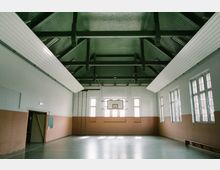 Innenansicht einer Turnhalle mit hoher Decke und sichtbaren Holzträgern. Die Wände sind teils weiß und teils holzfarben verkleidet, an der Stirnwand befinden sich ein Basketballkorb und große Fenster, die Tageslicht hereinlassen.