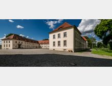 Weitläufiger, gepflasterter Innenhof mit beige verputzten Gebäuden im klassischen Stil, die rote Ziegeldächer haben. Umgeben von Bäumen und einer parkähnlichen Landschaft, zeigt der blaue Himmel mit wenigen Wolken eine sonnige Szenerie.