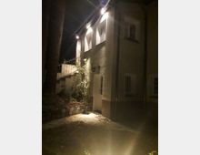 Zweistöckiges Wohnhaus bei Nacht mit gelblicher Putzfassade, beleuchtet durch Außenlampen an der Wand. Vor dem Gebäude befinden sich ein gepflasterter Weg, niedrige Pflanzen und ein kleines Steinbeet.