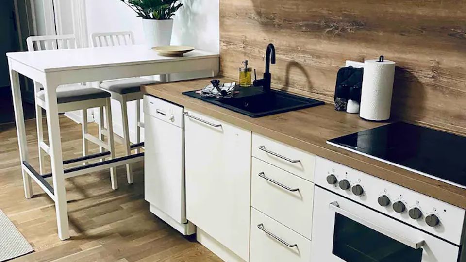 Helle, moderne Küche mit weißen Schränken, Holzarbeitsplatte und durchgehender Holzrückwand. Sichtbar sind Spüle mit schwarzer Armatur, Herd mit Backofen und Dunstabzugshaube sowie ein schmaler Esstisch mit hohen Stühlen auf Holzboden.