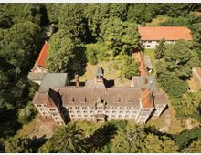 Luftaufnahme eines großen, historischen Gebäudes mit rotem Ziegeldach und symmetrischer Fassade, umgeben von dichtem Wald. Neben dem Hauptgebäude stehen weitere kleinere Gebäude mit ähnlicher Architektur im Grünen.