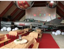 Ein Veranstaltungsraum in einer Halle mit einem silbernen Kampfflugzeug (Nummer 3046) als zentrales Dekorationselement, umgeben von Ballons und Lichtprojektionen. Im Vordergrund sind rustikale Holzbänke und Tische mit Jutedecken und Deko-Laternen auf roten Teppichen arrangiert.