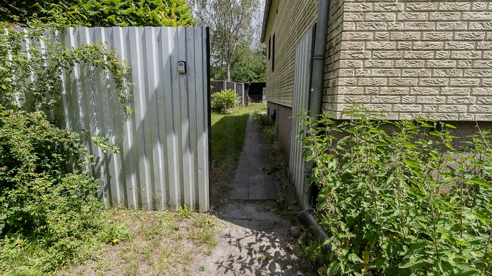 Schmaler Außenweg neben einem Wohnhaus mit heller Klinkerfassade, rechts mit Regenfallrohr und dichtem Unkrautbewuchs. Links steht ein hoher, grauer Wellblechzaun mit Eingang, dahinter Bäume und ein kleiner Gartenbereich.