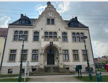 Historisches Gebäude mit heller Fassade, einer Uhr im Giebel und mehreren großen Fenstern mit Zierverzierungen. Vor dem Eingang führt eine Steintreppe hinauf, umgeben von Straßenlaternen und einem kleinen gepflasterten Vorplatz.