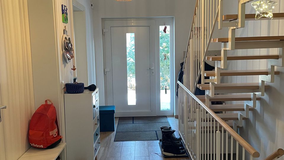 Heller Hausflur mit Holzoptik-Fliesenboden, weißer Eingangstür mit schmalen Glasfenstern und Deckenpendelleuchte. Rechts führt eine offene Treppe mit Holzstufen und weißem Geländer nach oben; links stehen eine Sitzbank und ein schmaler Schrank mit Garderobenbereich.