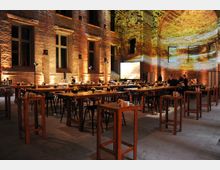 Innenraum einer historischen Backsteinhalle mit hohen Fenstern, stimmungsvoll beleuchtet durch warmes Licht. Mehrere hohe Holztische mit Hockern sind gleichmäßig angeordnet, dekoriert mit kleinen Blumenarrangements, während im Hintergrund ein großer Projektionsschirm sichtbar ist.