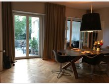 Esszimmer mit Parkettboden, modernem dunklem Esstisch und Stühlen sowie einer großen schwarzen Hängelampe. Im Hintergrund sind bodentiefe Fenster mit Blick auf eine Terrasse mit Pflanzen und Sitzmöbeln zu sehen.