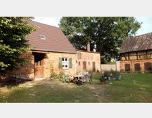 Backsteinhaus mit Satteldach in ländlicher Umgebung, ergänzt durch eine Scheune im Fachwerkstil und ein Nebengebäude aus Ziegeln. Im Vordergrund befindet sich eine Wiese mit Gartenmöbeln und einem Feuerkorb.