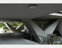 Überdachter Bereich eines modernen Gebäudes mit markanten, diagonal verlaufenden Betonpfeilern. Im Vordergrund befinden sich ein Fahrrad und niedrige Pflanzen, während im Hintergrund Tageslicht und Außenbereich sichtbar sind.