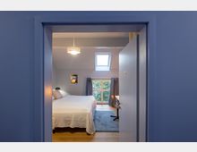 Blick durch eine geöffnete Tür auf ein modernes, schlicht eingerichtetes Schlafzimmer mit einem Doppelbett, einem Fenster mit Vorhängen, einem Oberlicht und einem kleinen Schreibtisch mit Stuhl. Der Raum hat helle Wände, Holzboden und eine minimalistische Deckenleuchte.