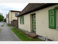 Eine Straßenansicht mit einem kleinen gelb gestrichenen Haus, das grüne Fensterläden und eine grüne Tür besitzt. Daneben verläuft ein gepflasterter Gehweg, und im Hintergrund sind weitere Gebäude und eine Ampel zu sehen.