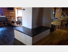 Gemütlicher Wohnbereich in einem Holzhaus mit sichtbaren Holzbohlenwänden, einer gemauerten Sitzecke mit dunklen Fliesen, Holzmöbeln und einem Boden aus Holz und dunklen Natursteinen. Im Hintergrund stehen ein weißer Sessel und ein Esstisch.