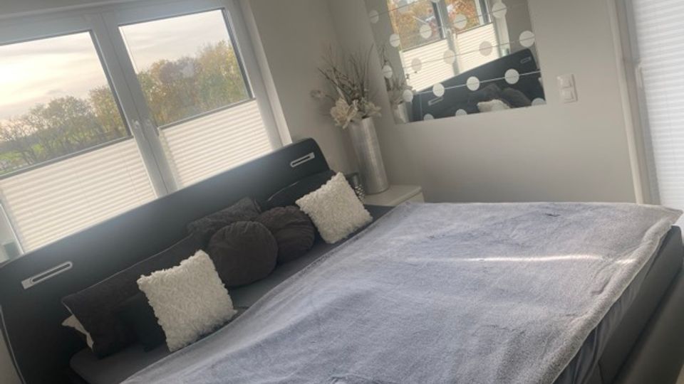 Schlafzimmer mit großem dunklem Polsterbett und grauer Tagesdecke, davor mehrere Kissen. An der Wand hängen ein Spiegel mit runden Lichtern, daneben stehen hohe Dekovasen; große Fenster mit Plissees lassen Tageslicht herein und geben den Blick auf Bäume frei.