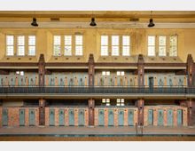 Historische Schwimmhalle mit zwei Ebenen von Kabinenreihen aus Backstein und bläulich-grünen Türen, darüber große Fenster mit farbigem Glas im oberen Bereich. Die Wände wirken gealtert, und Beleuchtung hängt von der hohen Decke.