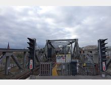 Metallbrücke mit Bahngleisen und Signalampeln, umgeben von modernen Gebäuden und einer Kirche mit einem spitzen Turm im Hintergrund. Die Brücke zeigt typische Stahlträgerkonstruktionen, und die Umgebung wirkt städtisch bei bedecktem Himmel.