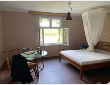 Ein schlichtes Schlafzimmer mit Holzboden, einem Doppelbett mit Baldachin, einem kleinen runden Tisch mit zwei Stühlen und einer offenen Fensterfront, durch die Bäume und Tageslicht sichtbar sind. Die Wände sind mit einem dezenten, gemusterten Tapetendesign gestaltet.