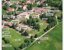Luftaufnahme eines ländlichen Freizeitgeländes mit mehreren Gebäuden und Grünflächen. Zu sehen sind beschriftete Bereiche wie ein Schloss, ein Landgasthof, ein Biergarten, ein Spielplatz, ein Park, eine Reithalle, sowie Wege und Wiesen, die zu Ausflügen einladen.
