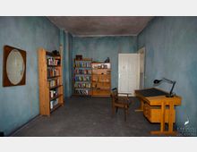 Ein einfacher Raum mit blauen Wänden und einem grauen Boden, ausgestattet mit zwei Holzregalen voller Bücher, einem Schreibtisch mit einer Lampe, einem Stuhl und einem ovalen Spiegel an der Wand. Im Hintergrund befindet sich eine weiße Doppeltür.