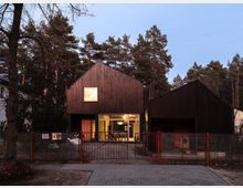 Moderne Wohngebäude mit zwei dunklen Giebelhäusern aus Holz, getrennt durch einen Glasbereich mit großen Fenstern, die einen Einblick ins Innere ermöglichen. Im Vordergrund befindet sich ein Metallzaun mit Tor, umgeben von einer Waldlandschaft mit hohen Kiefern.