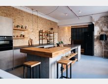 Moderne Küche mit industriellem Design, gekennzeichnet durch eine roh belassene Backsteinwand, offene Regale und eine Kücheninsel mit Holzplatte. Die Einrichtung umfasst eine schwarze Spüle, Hocker mit Holzsitzen und Details wie Glaspendelleuchten und Metallakzente.