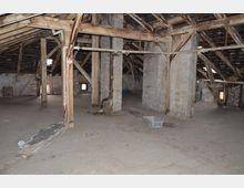 Dachboden eines alten Gebäudes mit freiliegenden Holzständern, schrägen Dachbalken und unverputzten Wänden. Der Boden ist staubig, und es sind teils Bauschutt und ein Plastikkasten verteilt.