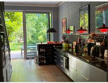 Moderne Küche mit langen schwarzen Arbeitsflächen, Edelstahlspüle und Schränken an einer grauen Ziegelwand mit Postern und roten Pendelleuchten. Große Glastüren führen auf eine Terrasse und in einen grünen Garten.