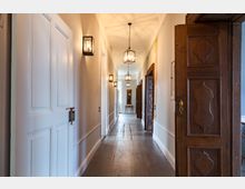 Langer, heller Flur in einem historischen Gebäude mit Holzböden, weißen Wänden und hölzernen Türen. An der Decke und den Wänden hängen Laternenlampen, und am Ende des Flurs steht ein kleiner Tisch mit einem Spiegel darüber.