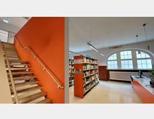 Innenansicht einer modernen Bibliothek mit einer offenen Treppe auf der linken Seite vor einer orangefarbenen Wand. Rechts befinden sich Bücherregale, ein Schreibtisch mit Computer und große Fenster mit Rundbögen, die viel Tageslicht hereinlassen.