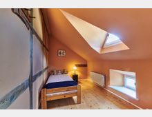 Dachzimmer mit einer schrägen, orangefarbenen Decke, einem Holzbett mit Stofftieren, einer kleinen Nachttischlampe, einem Heizkörper unter einem kleinen Fenster und einem Dachfenster, das Licht hereinlässt. Die Wände sind teilweise mit sichtbaren Holzbalken verziert.
