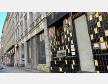 Straßenansicht mit einer Reihe von Altbauten, darunter eine Fassade mit schwarzen und gelben Kacheln, geschlossenen Rollläden und dekorativen Elementen wie einem Regenbogenbanner. Andere Gebäudefronten zeigen verglaste Fenster mit Metallgittern und vereinzeltem Graffiti.