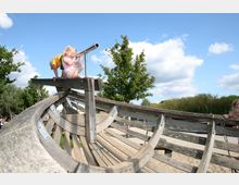 Zwei Kinder spielen auf einem hölzernen Spielgerät in Form eines Schiffes in einem Outdoor-Spielplatz. Im Hintergrund sind Bäume, ein sandiger Boden und ein Gewässer unter einem blauen Himmel mit einigen Wolken zu sehen.