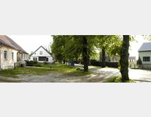 Straßenszene in einer ländlichen Umgebung mit bäuerlichen Häusern, darunter ein weißes Fachwerkhaus mit Zickzack-Giebel und ein helles Wohnhaus mit roten Ziegeln. Die Straße wird von hohen Laubbäumen gesäumt, und im Hintergrund ist eine kleine Steinmauer mit Gebäuden erkennbar.