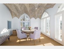 Heller Raum mit gewölbter Decke, die mit floralen Mustern verziert ist, großen weißen Rundbogenfenstern und einer Sitzgruppe mit rundem Holztisch und lila gepolsterten Stühlen. Der Raum ist mit hellem Teppichboden ausgelegt und bietet Blick in eine Gartenlandschaft.