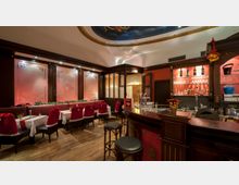 Elegantes Restaurant mit festlicher Dekoration, einschließlich Stühlen mit roten Überzügen und Tischdecken sowie weihnachtlichem Schmuck. Im Vordergrund eine Holztheke mit Barhockern, im Hintergrund dekorative Glaswände mit Muster und Regale mit Flaschen und Gläsern.