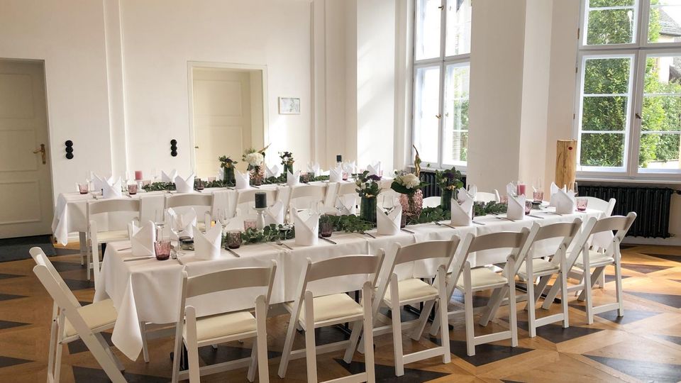 Heller, hoher Festsaal mit weißen Wandpaneelen und großen Sprossenfenstern, durch die Tageslicht und Blick ins Grüne fällt. Zwei lange Tafeln mit weißen Tischdecken, gefalteten Servietten, Blumen- und Kerzendekoration stehen auf einem gemusterten Holzparkettboden.