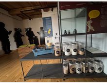 Ein Ausstellungsraum mit Holzfußboden und Holzbalkendecke, in dem verschiedene Gläser mit Getreide und Mehl auf einem Regalsystem stehen. Im Hintergrund sind Silhouetten von Menschenfiguren an einer Wand sowie historische Informationen und eine Waage zu sehen.