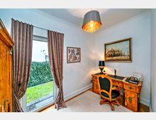 Ein kleiner Arbeitsraum mit antikem Holzschreibtisch und passendem Stuhl. An der Wand hängen zwei Bilder, darunter eine Stadtansicht. Links führt eine Glastür mit dekorativen Vorhängen in einen Garten.